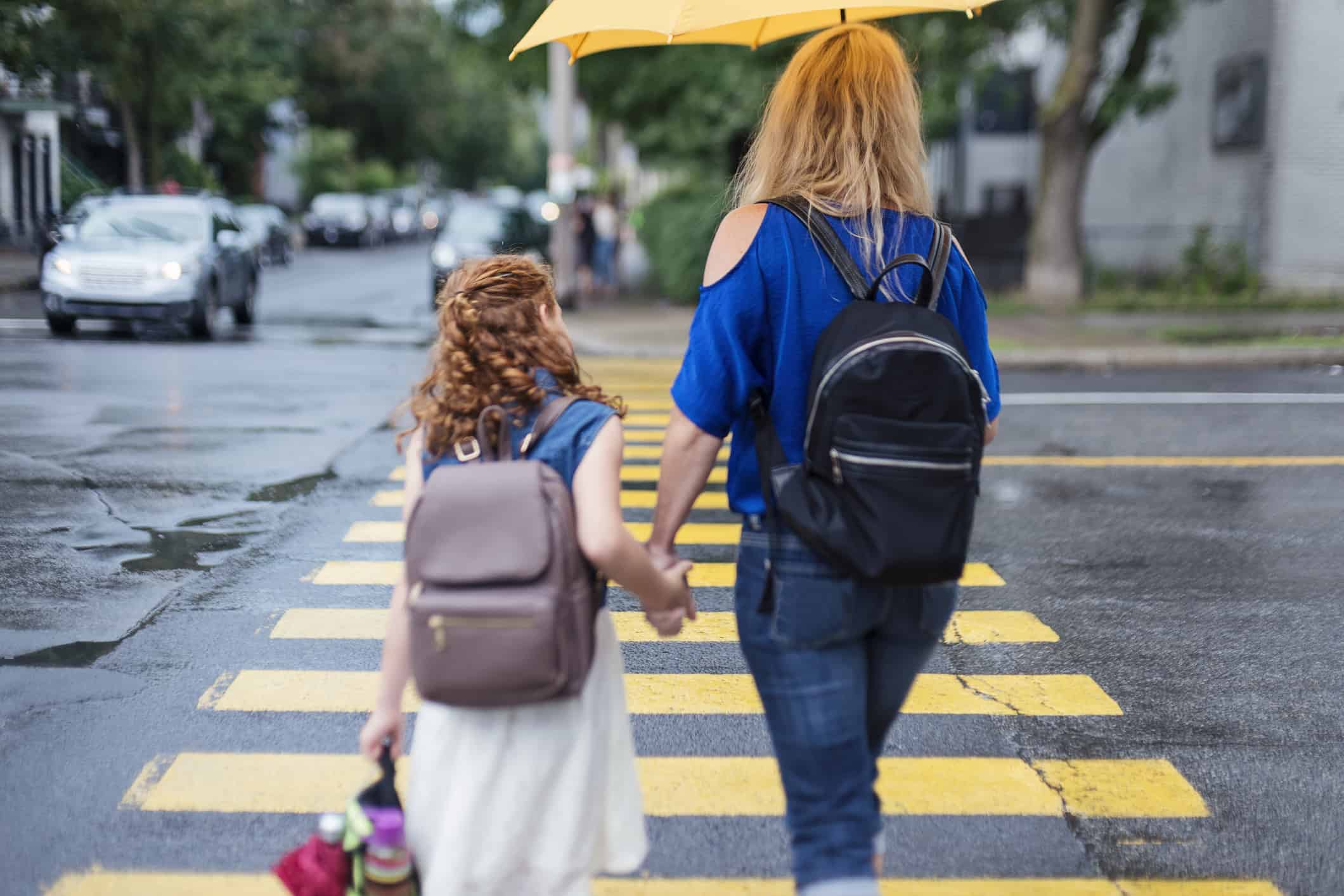 Young redhead girl and her single mother walking to school. Back view of them holding hands, crossing city street on rainy day. Horizontal waist up outdoors shot with copy space. This is part of a « Day in the life of a child » serie and was taken in Montreal, Canada.