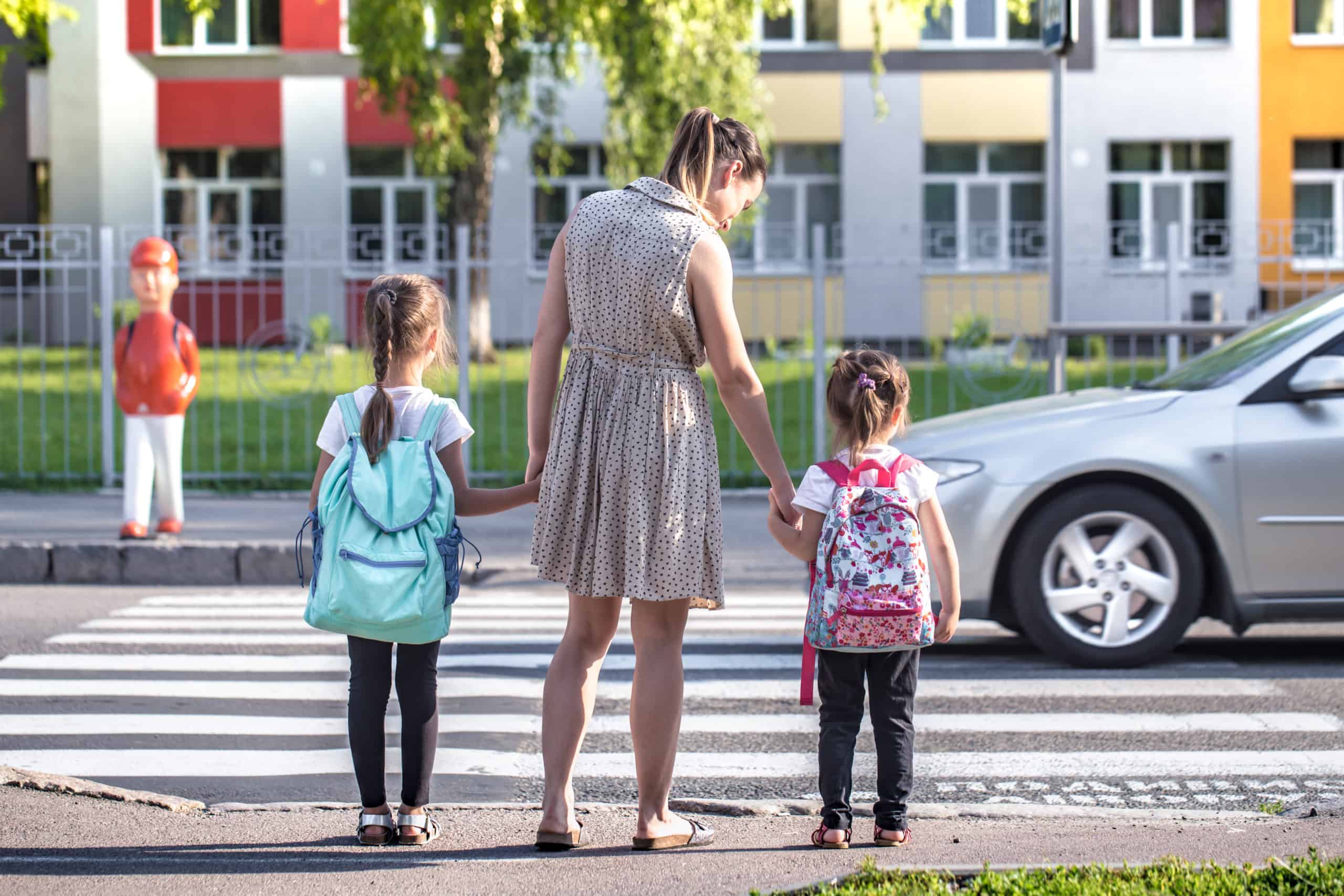 Mother holding both girl kids with backpacks hands waiting at a crosswalk